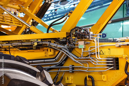 Agricultural harvester hydraulic control valve block steel tubes. Modern farming machinery fluid power manifold component on yellow frame chassis with technical engineering vehicle details.