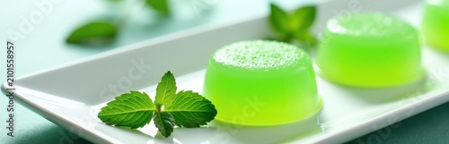 Vibrant green gelatin desserts sit on a white rectangular plate garnished with fresh mint leaves. These translucent sweets offer a refreshing treat perfect for summer occasions and light meals.