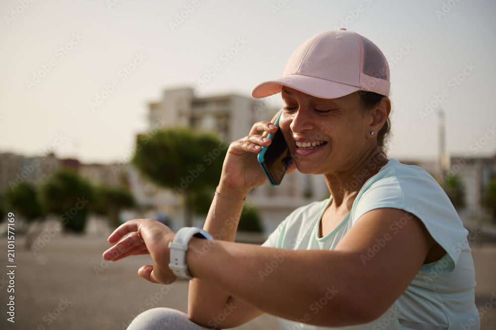 Obraz premium Smiling woman checking smartwatch while talking on phone during outdoor fitness break