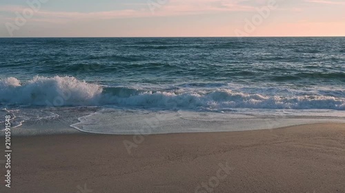 Gentle ocean waves roll onto the sandy beach as the warm golden light of the setting sun reflects beautifully across the water’s surface