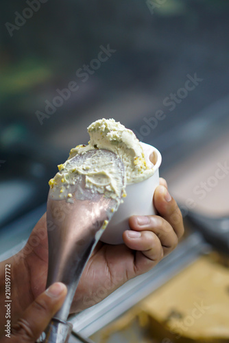 Fresh artisan gelato served into a white paper cup using a metal scoop. Authentic preparation process with shallow depth of field, ideal for hospitality, dessert and food service concepts.