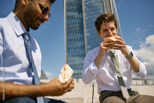 Φωτογραφία Indian and Caucasian young adult businessmen eating sandwiches during outdoor lunch break near office tower, representing workplace routine