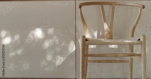 Sunlight streams through a window, casting dappled shadows on a minimalist kitchen's sleek white cabinets. A simple, elegant wooden chair with a woven seat sits in the foreground,