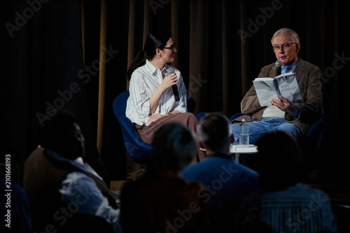 фотографія Wide shot of senior Caucasian man reading from book while young woman moderating stage discussion before audience