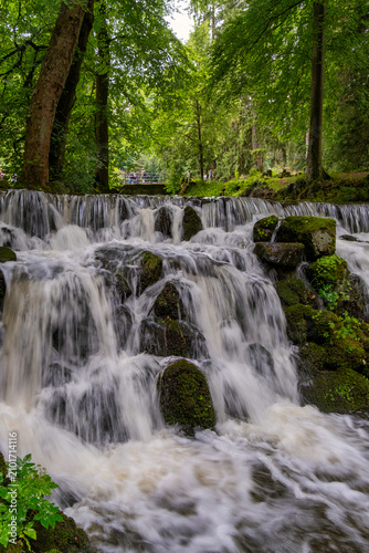Tranquil Waterfall With Lush Surroundings, Serene Waterfall Flowing Beneath Stone Bridge Amidst Vibrant Woods