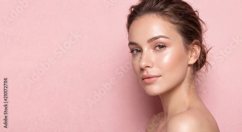 Side profile portrait of a beautiful young Caucasian woman with clean glowing skin and hair tied up looking over her shoulder against a solid pastel pink background