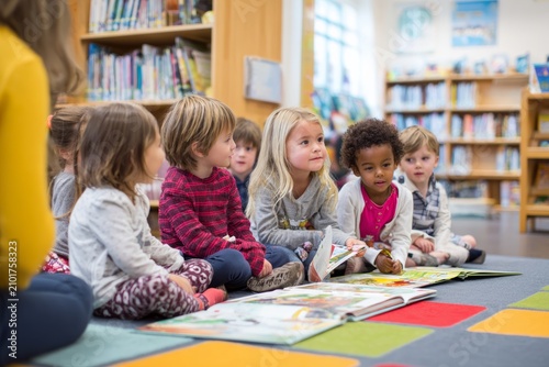 back to school: children sitting on colorful rug in reading corner library, flipping through picture books together. Teacher or librarian nearby, bookshelves, warm and inviting classroom environment. 
