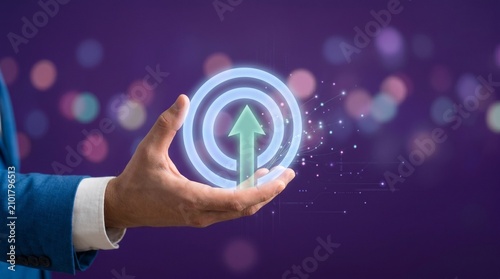 Hand holding a glowing circular target icon with an upward green arrow symbolizing growth and success against a blurred purple background