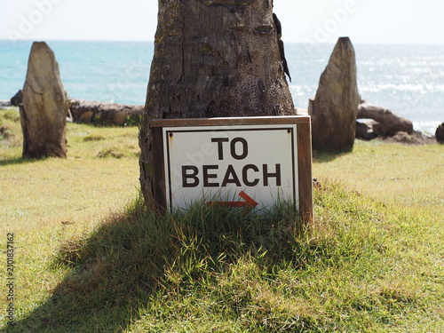 Sign showing direction to beach near ocean and grass area on sunny day.