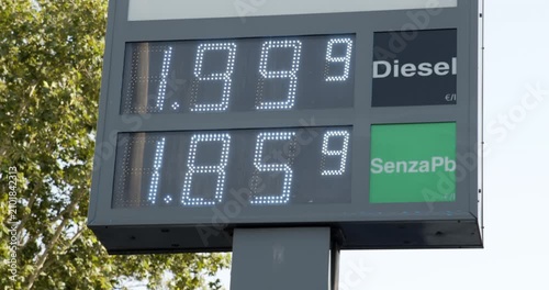 A close-up shot of a digital gas station price sign showing fuel rates for diesel and unleaded petrol, with green tree foliage partially visible in the blurred background under daylight.