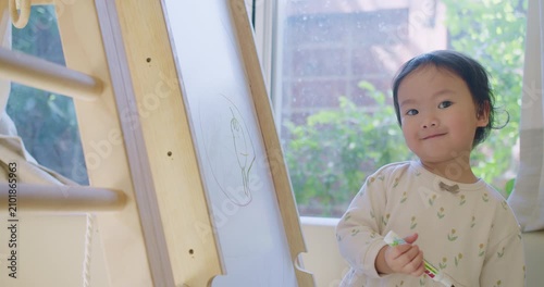 A cute toddler girl proudly smiles at the camera while drawing on a whiteboard easel, engaging in creative play for her early childhood development and homeschooling.