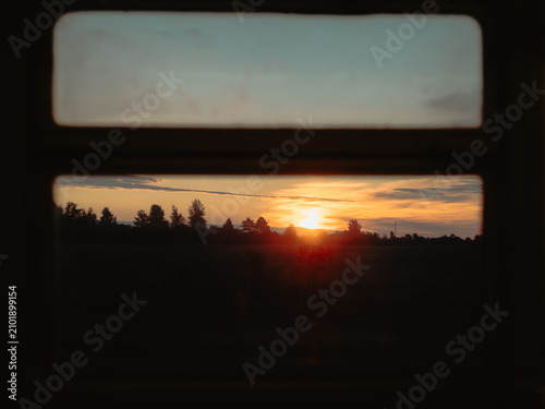 Golden sunset over a dark forest and field silhouette, framed by a retro train or bus window during a evening trip.