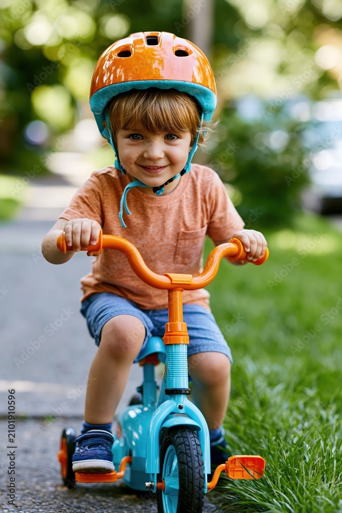 Obraz premium Smiling child riding tricycle on sidewalk wearing helmet and summer clothes