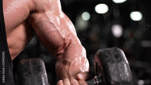 Close-up of a flexed bicep at peak contraction during a dumbbell curl, veins and muscle striations visible under gym light. Muscle definition workout concept