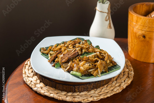 Принти на полотні Savory Noodle Dish on Banana Leaf: A delicious and expertly prepared noodle dish served on a fresh banana leaf, presented on a white plate atop a rustic wooden table, hinting at a culinary delight