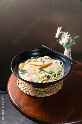 Konstfotografi Spicy Noodle Soup: A vibrant bowl of spicy noodle soup, brimming with an assortment of fresh ingredients, served with chopsticks on a rustic wooden table, promising a flavorful culinary experience