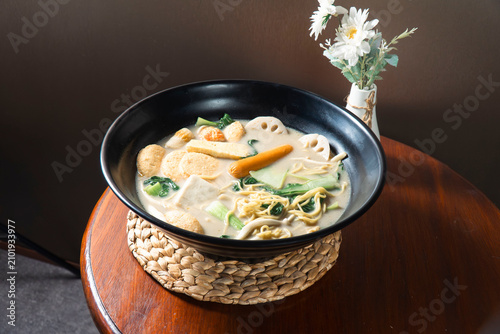 Foto Hearty Noodle Soup: A steaming bowl of flavorful noodle soup, brimming with an assortment of fresh vegetables, tofu, and tender noodles, served in a dark ceramic bowl atop a woven placemat
