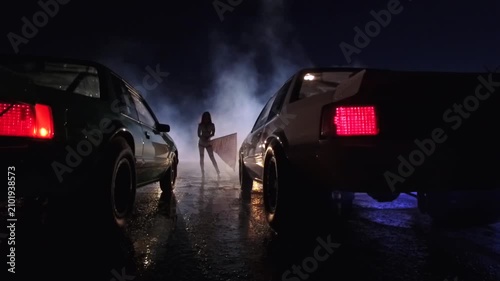 Street racing scene at night with a woman holding a checkered flag between two muscle cars