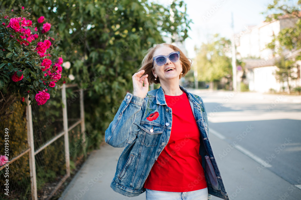Fototapeta premium Laughing mature woman 55-60 year old wearing sun glasses and denim jacket outdoors over flowers close up. Looking at camera.