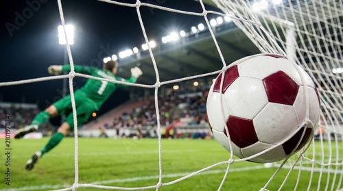 Classic white and dark red soccer ball hits the white mesh net while an agile goalkeeper dives in the background during a professional night match under bright stadium floodlight illumination.