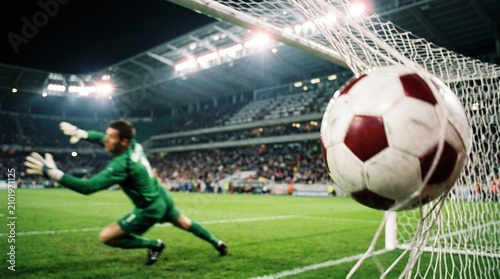 Professional soccer goalkeeper in a green uniform dives to block a fast moving ball that is hitting the back of the net during a high stakes night time stadium match