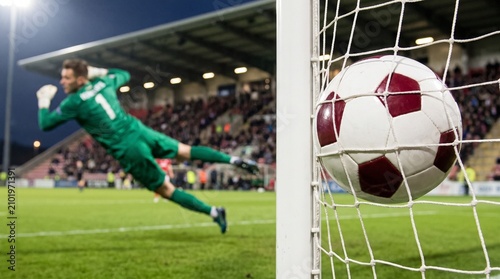 Soccer ball hitting the back of the net after a goal is scored while a professional goalkeeper dives in the background during a competitive evening match under bright stadium lights