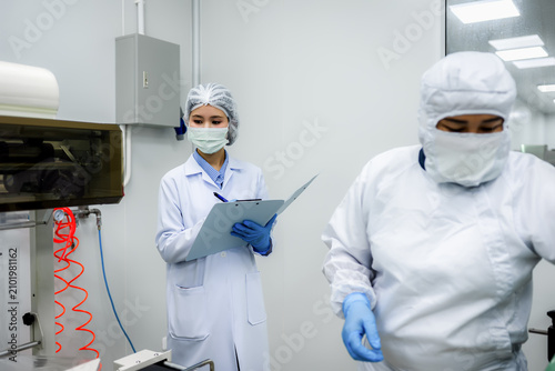 Female quality control inspector wearing sterile uniform auditing pharmaceutical production line. Professional team workspace scene for cosmetic biotechnology industry quality assurance.