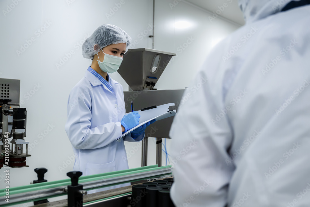 Naklejka premium Asian woman scientist in sterile uniform checking pharmaceutical product process on conveyor, concept of hygiene and quality assurance management.