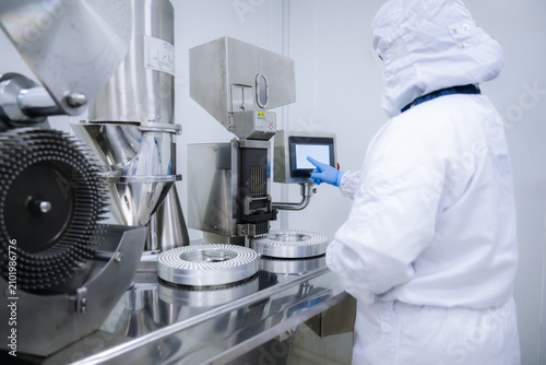 Pharmaceutical worker using medicine control digital panel in factory. Operating capsule filling machine via touchscreen in clean room. Pharmaceutical manufacturing with touchscreen interface.