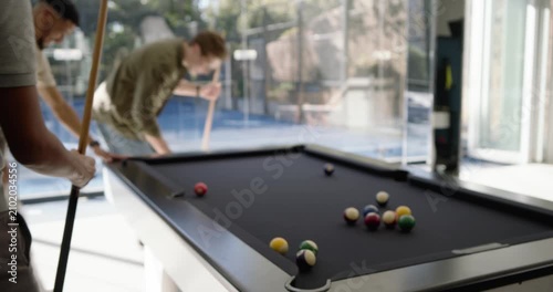 Diverse male friends in glass room playing pool, cue striking and scattering balls on black table