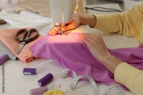 Woman sewing fabric with professional machine at white table in workshop, closeup