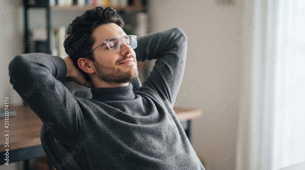 Obraz premium Satisfied young male freelancer wearing glasses relaxing in comfortable chair with hands behind head and closed eyes enjoying peaceful break at home office after finishing successful project.