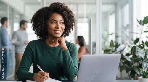 Smiling black businesswoman with curly afro hair writing in notebook near laptop at modern office, happy African American female manager working at desk, corporate professional employee.