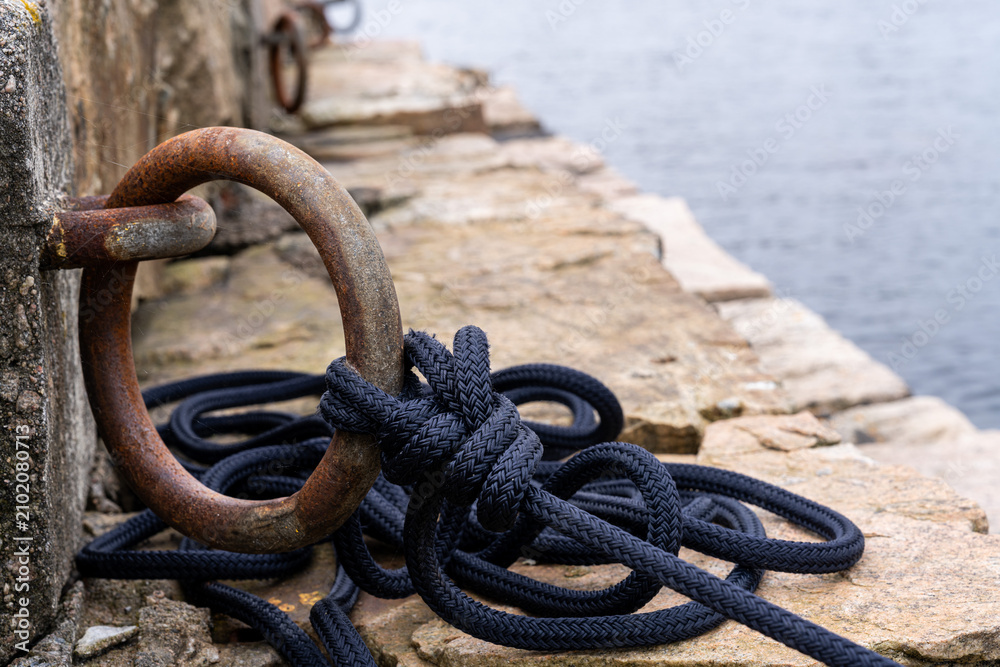 Obraz premium Rusty mooring ring with black rope on stone pier
