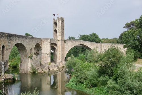 Besalu, Spain - June 16, 2026: View of the medieval Pont Vell (Old Bridge) over the Fluvia river, with its central fortified gate tower flying flags above the stone arches, Girona province, Catalonia.