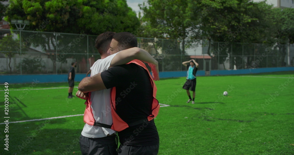 Obraz premium Soccer teammates embrace and raise fists in celebration after scoring goal on soccer field, shouting with joy, while kneeling opponent watches, showing unity