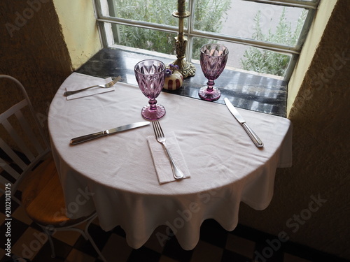 A round table is prepared for a meal at a small restaurant. There are two purple glasses, forks, knives, and napkins. The light shines through the window.