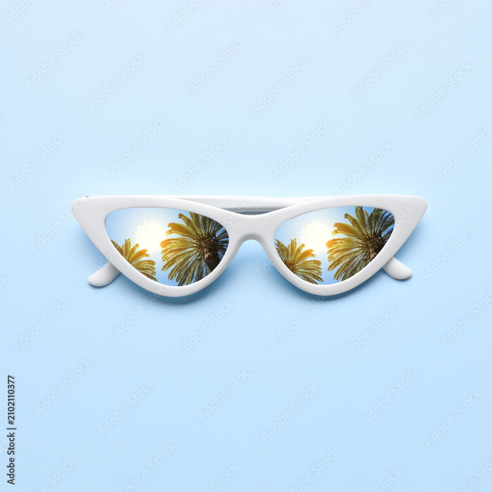 Fototapeta premium Stylish sunglasses on light blue background. Sky and tropical palm trees reflecting in lenses