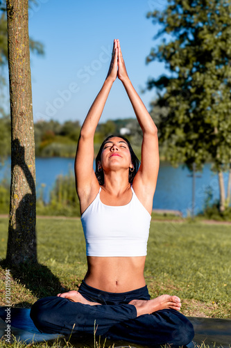 A young Hispanic woman practises a seated pose with arms overhead in a peaceful park by the lakeside in summer, nurturing wellness and a positive mindset by the water.