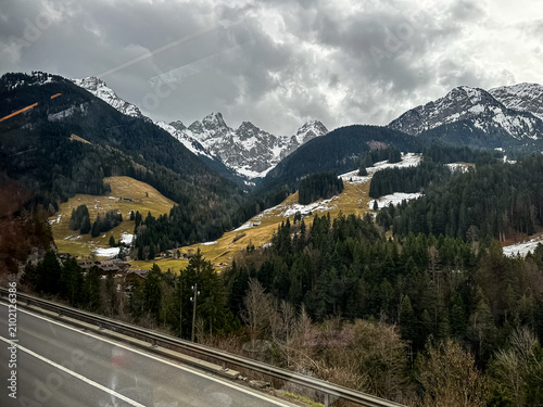 A dramatic alpine landscape highlights towering, jagged mountain peaks covered in patches of snow under a heavy, overcast winter sky. Dense green pine and fir forests blanket the steep valley walls, s