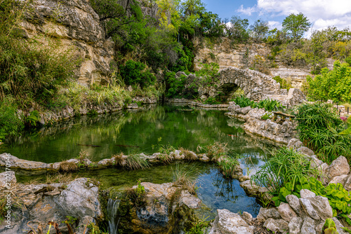 Picturesque stone arch bridge over a clear pond with a small waterfall and lush green plants against limestone cliffs in San Antonio, Texas.