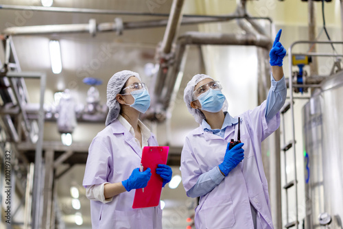 Female engineers inspecting metal storage tank and piping system in fish can food factory, Quality control specialists auditing production standards large stainless steel vats in industrial plant.
