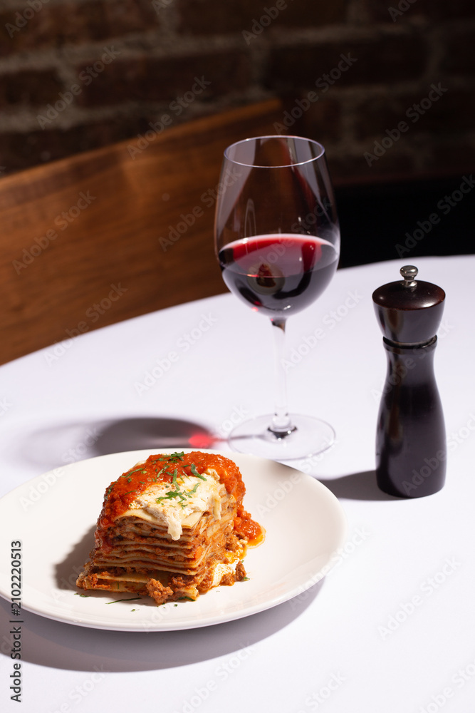 Naklejka premium Angled shot of beef lasagna slice with a glass of red wine and pepper mill, restaurant table setting
