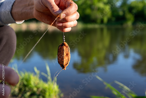 Quadro su tela Close-up of a fisherman holding a method feeder packed with groundbait prepared for carp fishing beside a pond