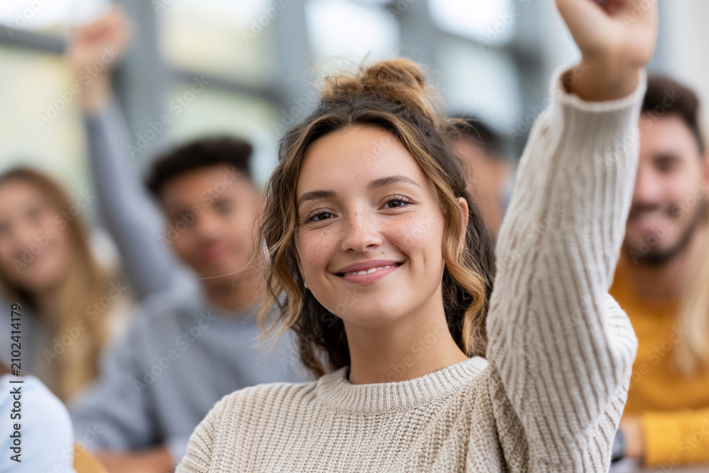Obraz premium Young woman smiles, raising hand in classroom. Group of students behind her shows engagement. Her expression conveys enthusiasm and confidence. Ideal for education, learning