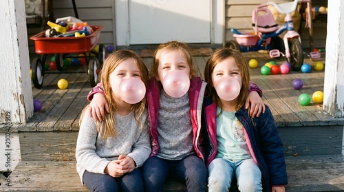 Three sisters blowing pink bubblegum bubbles together on porch