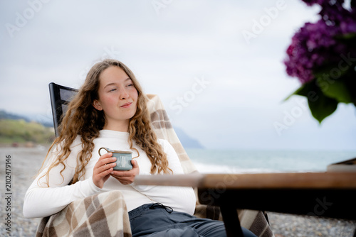 Принти на полотні Portrait of a peaceful young woman enjoying a quiet moment with a warm drink on the beach during the day, enjoying solitude and practicing reflection in nature, and slow living by the sea
