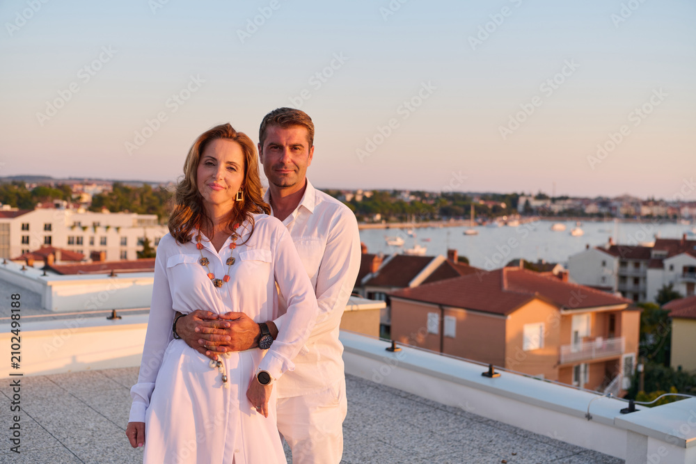 Obraz premium Middle-Aged Couple Embracing on Rooftop Terrace With Seaside View at Sunset Overlooking Marina and Town