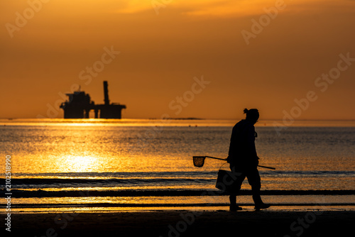 Clam digger carrying a bucket and net at dawn, industrial sea platform silhouette, contrast concept.