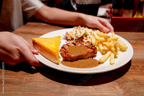 Hands holding a plate of grilled pork chop steak with black pepper gravy, crispy french fries, fresh coleslaw, and buttery toast on a wooden restaurant table. Delicious casual dining meal concept.